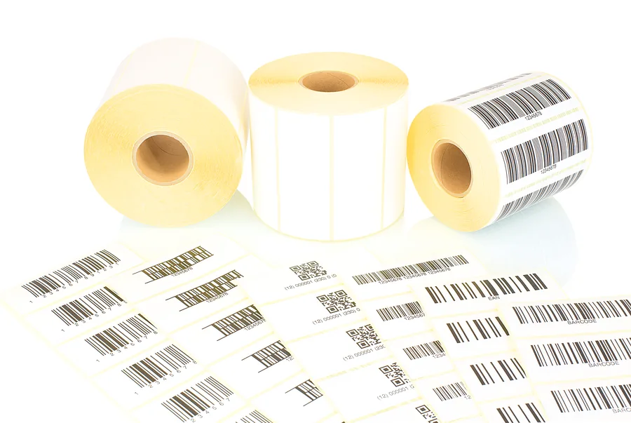 Trois rouleaux d’étiquettes blanches sur mandrin carton, l’un portant des codes-barres, devant des étiquettes à codes-barres et QR codes